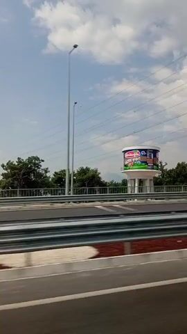 A video captures a view from a moving vehicle, likely a car, traveling on a highway. In the background, a large cylindrical water tower stands prominently, adorned with a colorful advertisement. The advertisement features images of what appear to be food products, including a container with a red lid and colorful labels with text in an unfamiliar language. The tower itself is supported by a white, tiered structure with visible struts. Beyond the tower, a line of trees and distant hills are visible under a partly cloudy sky. Overhead, power lines stretch across the frame, supported by tall poles, including a tall lattice structure. A black car is seen in motion on the highway in the middle ground, creating a slight motion blur. The foreground shows the highway lanes, with a barrier made of metal railings and brick-like material separating the lanes. The camera angle suggests a slightly elevated perspective, looking forward and to the side. The overall impression is a roadside view of infrastructure and advertising in a daytime setting.