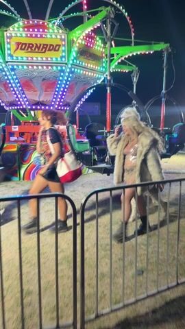 At an outdoor night carnival, two women are walking near a ride labeled "Tornado" with lights. The woman in front is wearing a faux fur coat over a sparkly top and lace skirt. She stops, smiles, and waves to the camera. The other woman wears a crown. A person walking behind the two women carries a red and white tote bag. The camera blurs.