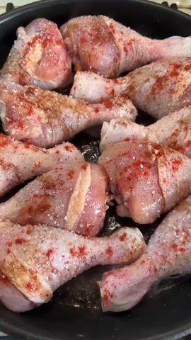 This media file shows a close-up, overhead view of raw chicken drumsticks being seasoned and cooked in a black frying pan. The chicken pieces are arranged closely together, covering most of the pan's surface. They are sprinkled with salt and a reddish spice, likely paprika or chili powder, and some darker spices, possibly black pepper or herbs. Some of the chicken pieces are beginning to sear, indicated by a slight browning and the visible steam rising from the pan, suggesting they are actively cooking. The lighting is bright, suggesting it could be daytime, and there are no discernible external environmental cues to indicate the specific location beyond the cooking activity. The focus is entirely on the food preparation process. There is no visible text in the media file.