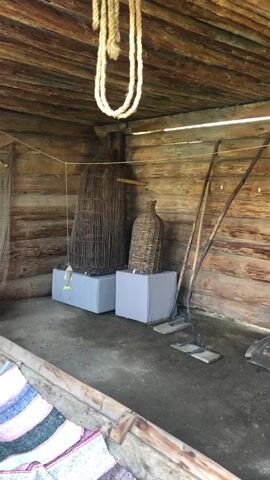 The video shows the interior of a rustic wooden structure, possibly a shed or boathouse. The walls and ceiling are constructed from rough-hewn logs. In the foreground, a portion of a woven mat or rug is visible. The middle ground features several items that suggest a connection to fishing or water activities. Two large wicker baskets, one tall and conical, the other shaped like a bottle, are displayed on grey pedestals. To the right, two long-handled tools, resembling hoes or rakes, lean against the wall. Suspended from the ceiling is a thick rope. The back wall displays various fishing nets of different sizes and weaves, as well as a circular net or trap. A wooden boat, weathered and possibly burnt, rests on supports in the far right of the frame. A small, white, fish-shaped object hangs from a string in the center of the back wall. The floor is dirt or packed earth. The overall impression is of a historical or ethnographic exhibit showcasing tools and artifacts related to traditional livelihoods.