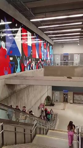 The video shows an indoor scene with people moving about in a modern transit station. Several individuals are going down a set of stairs, and some are on an escalator going down. The walls are concrete, with a large, colorful mural on one side. The mural has geometric shapes and figures depicting people cheering. The ceiling is a grid of industrial lights. An upper level of the station is visible, with enclosed rooms and doors. There's a sign with an arrow indicating direction. The overall environment suggests a busy public space.