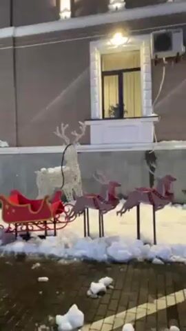 The video shows a Christmas-themed outdoor display set up outside a building with visible snow on the ground. The decorations include a red and gold sleigh, reindeer figures, and several wooden structures decorated with stars. The building behind the display is a multi-story structure with multiple windows, illuminated by interior and exterior lights. The overall scene suggests a festive atmosphere, potentially for a Christmas market or event. The camera appears to be moving slowly.