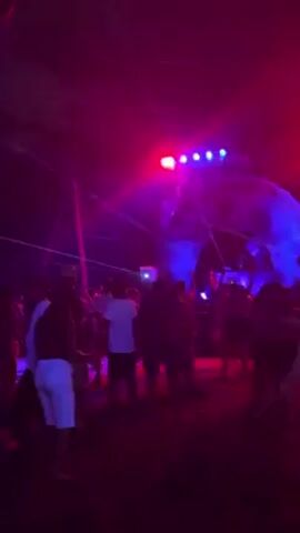 The video shows a crowd of people gathered under a tent, illuminated by colorful lights and laser beams.  Many people are wearing white shirts and dark shorts. The tent has a design projected onto it.  A thatched-roof bar is visible in the background.  The overall atmosphere suggests a party or outdoor event with music and dancing.