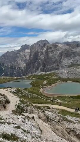 The video shows a mountainous landscape with two lakes. There are people walking on a path near the lakes. In the background, there are tall, jagged mountains. The sky is cloudy.