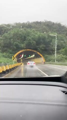 A white car drives into a tunnel on a rainy day. The road is wet and reflects the lights. Yellow traffic cones line the sides of the road. The tunnel entrance is arched and illuminated by yellow and black stripes. Traffic lights inside the tunnel are visible. The view is from inside another vehicle, looking through the windshield.