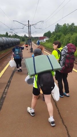 Two people are standing on a train platform with large backpacks and camping mats. The person in the foreground is wearing a green and yellow jacket and black shorts, carrying a large black backpack and a white plastic bag. They have a green sleeping mat strapped to their backpack. Behind them, another person in a blue jacket and brown pants carries a white backpack and a white plastic bag. The person further back wears a bright yellow shirt and black pants with a large maroon and grey backpack. They are all facing away from the camera and appear to be waiting for a train.

The scene appears to be at a train station, with train tracks and overhead electrical wires visible. A train is parked on the tracks on the left side of the frame. The platform is made of concrete with yellow lines marking the edge. Trees and green foliage are visible in the background, suggesting an outdoor setting. The sky is overcast, indicating a cloudy or potentially rainy day. The time of day is difficult to determine precisely, but it appears to be daytime. The overall mood is one of travel and preparation for an outdoor adventure.

No visible text is present in the image. The location context provided is "Орехово, Russia," which helps to place the scene geographically.
