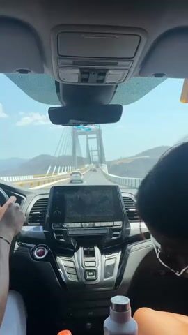 The video provides a first-person view from inside a car, likely a Honda Odyssey, as it drives onto and across a large cable-stayed bridge. The time of day is bright and sunny, with a clear blue sky and some white clouds. Mountains are visible in the distance on either side of the bridge.

In the foreground, a person's left hand is visible on the steering wheel, indicating they are driving. They wear a small, beaded bracelet. The car's dashboard is prominent, featuring a central infotainment screen that appears to display a map or navigation. Below the screen are various controls and a push-button gear selector with "D/S" visible. Two bottles are placed in cup holders in the center console: one plastic bottle containing an orange liquid, and a white or silver metal water bottle. The partial head of another person (likely a passenger) is visible on the far right of the frame.

As the car moves onto the bridge, its impressive white pylons and numerous suspension cables become the dominant feature of the view through the windshield. Other vehicles can be seen ahead on the bridge. The audio includes speech and an exclamation, suggesting excitement or awe at the scenery or the bridge itself. The setting is consistent with a scenic drive in a location such as Tula del Río, Mexico.