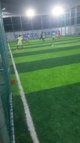 A high-angle static shot from behind a green net shows an indoor soccer field with bright green and dark green striped artificial turf. Above the white sideline, approximately a dozen people are playing soccer. Most players wear short-sleeved shirts and shorts, in various colors including white, yellow, and pink. Many bright lights illuminate the field from above, creating shadows. In the background, a white wall with the words "DAKATA ARENA" in blue letters is visible.