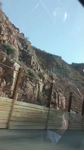 The video shows a view from inside a vehicle, looking out at a steep, rocky mountainside. A retaining wall constructed of horizontal wooden planks supported by vertical posts lines the side of the road, obscuring the lower part of the mountain. Above the retaining wall, the rocky slope is visible, with some areas appearing to have been reinforced with metal netting and support structures. The sky is clear and blue. The vehicle appears to be moving, as indicated by the slight blur and the changing perspective. There are no people or other notable objects visible in the scene.