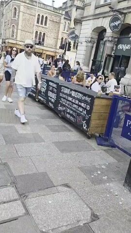 This media file shows a street scene in what appears to be a busy urban area. The primary subjects are people walking on a paved sidewalk. In the foreground, a man with a beard, wearing a hat, sunglasses, a white short-sleeved shirt, and distressed denim shorts, walks towards the camera. He has tattoos on his arm. Behind him, several other people are walking in the same direction.

To the right of the frame, there is a long, black structure with wheels that appears to be a mobile bar or seating area for an establishment. This structure has various types of text written on it. It displays information about "FRIDAY NIGHTS KARAOKE DJ," "SATURDAYS LIVE MUSIC FROM 7PM DJ BYRON & GUINESS," and "MONDAYS QUIZ NIGHT FREE ENTRY / HAPPY HOUR - MON-FRI 5PM - 8PM."

In the background, there are buildings, including one with a sign that reads "O'NEILLS." There are also streetlights and what appear to be other pedestrians and patrons of the establishment. The scene suggests a daytime setting, possibly late afternoon or early evening, given the lighting. The weather appears to be overcast but pleasant enough for people to be out and about. The overall atmosphere seems lively and social, with people enjoying a day out in what is identifiable as Cardiff, United Kingdom, based on the provided location context.