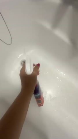 A first-person perspective video shows a hand spraying a white bathtub with a cleaning product. The hand is holding a spray bottle with a white nozzle and a purple and orange bottle. The spray is visible in the air and on the surface of the bathtub. The bathtub is white and appears to be made of porcelain or acrylic. A drain is visible in the bottom of the bathtub. The video is lit by overhead lighting, casting shadows on the bathtub. The overall scene is clean and well-maintained.