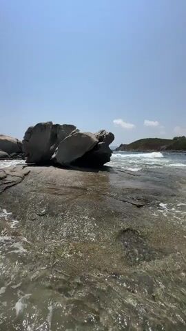 The video shows a beach with rocks and the ocean. The water is clear and shallow, with waves gently lapping against the shore. Large, grey rocks are scattered across the beach, some of them smooth and rounded, others jagged and angular. The sky is a clear blue with a few wispy clouds. The sun is shining brightly, casting long shadows across the sand. The overall scene is one of natural beauty and tranquility.