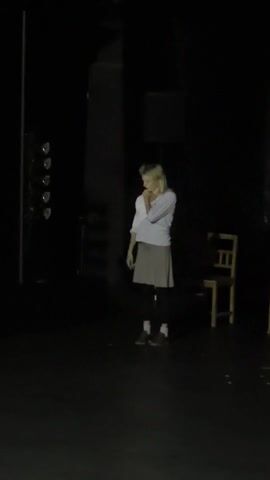 A woman with blond hair, wearing a white shirt and gray skirt, stands alone in a dark room under a spotlight. Her hands are near her face, and she appears to be speaking or signing. To her left, there is a lighting rig with four lights. To her right, there are two wooden chairs. The floor is dark and reflective. The background is entirely black.