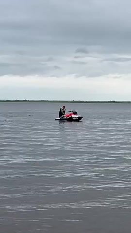 This video shows a person riding a jet ski on a large body of water. The sky is overcast with dark, cloudy conditions, suggesting it might be an early morning or late afternoon, or simply a gloomy day. The water appears calm with gentle ripples. In the background, a thin strip of land with greenery is visible on the horizon, indicating a shoreline. The jet ski is red and black, and the rider is wearing a helmet and a life vest, suggesting safety precautions are being taken. The overall scene is one of recreational activity on the water under a gray sky. The timestamps indicate a short duration, showing the jet ski moving slightly across the frame. There is no visible text in the video.
