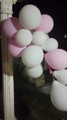 A bunch of pink and white balloons are hanging from a string against a dark background. The balloons are of various sizes and shapes. Some are round, while others are elongated. The string is attached to a white column on the left side of the frame. In the background, there is a hint of greenery and what appears to be a house or building. The lighting is dim, suggesting it is nighttime or dusk.