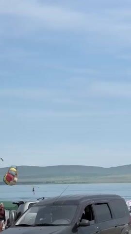 This is a vertical, medium shot of a parasailer being towed over a body of water. The parasail is multicolored, with red, yellow, blue, and white panels. A person is suspended beneath the parasail by a harness and rope. The water is calm and a shoreline with low, rolling hills can be seen in the background. In the foreground, there are several parked vehicles. One is a dark-colored SUV or minivan, partially visible on the left side of the frame. Another is a silver car with its trunk open, loaded with luggage, parked to the right of the first. To the far right, a portion of a beige tent is visible. The sky is blue with scattered white clouds. The scene appears to be a recreational area, possibly a beach or lakefront, on a clear day.