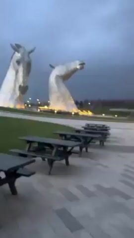 The video shows two large, metal horse head sculptures, likely the Kelpies in Scotland, standing on a green lawn under a cloudy, grey sky. Several dark-colored picnic tables are lined up on a paved area in the foreground. The sculptures are illuminated, and their surfaces appear to be composed of many small, reflective pieces. A few people are visible in the distance near the base of the sculptures.