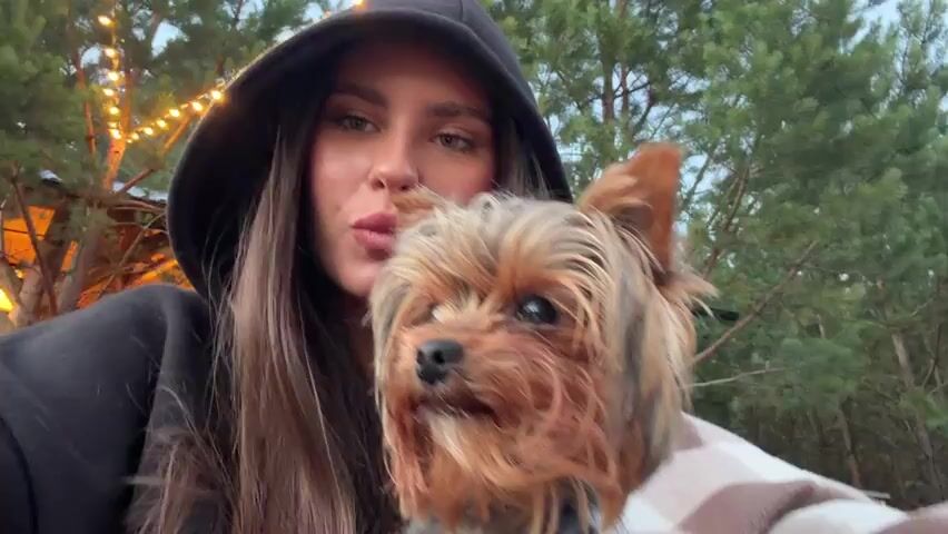 A woman wearing a black hoodie with the hood up is holding a small Yorkshire Terrier dog. The dog is wearing a brown harness. The woman is looking at the camera and making kissing faces and smiling. The dog is looking slightly to the side. They are outdoors, with trees and string lights visible in the background. The overall lighting suggests it is dusk or evening. There is a wooden structure with lights in the background.