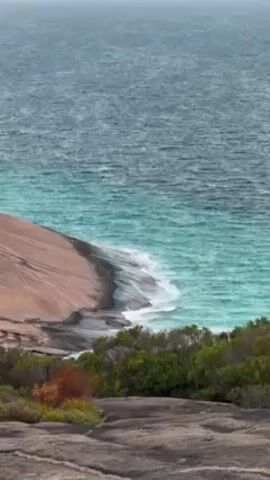 The video shows a wide shot of the ocean, with a rocky coastline in the foreground. The water is a mix of turquoise and dark blue, with small waves crashing against the shore. In the distance, there are several islands visible. A small boat is seen moving across the water, leaving a white wake behind it. The sky is overcast with clouds.