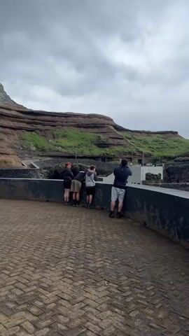 The video shows a group of five people standing at a viewing platform overlooking a landscape. The platform has a low concrete wall, and the ground is paved with a herringbone pattern of bricks. Behind the group is a layered rock formation with some vegetation growing on it. The sky is overcast. 

Another two people, a man and a woman, appear from behind the rock formation and walk towards the group. They are walking on a paved path that leads to the viewing platform. The man is wearing a patterned pink shirt and white pants, while the woman is wearing a white top.