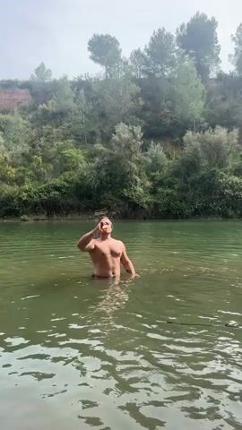 Here is a description of the video:
A man stands waist-deep in a calm, green river, surrounded by lush greenery. He is shirtless with dark hair pulled back and has a bottle of Corona beer in his hand. He smiles and makes a peace sign with his other hand. The background consists of trees and a hillside with vegetation. The lighting suggests a bright, slightly overcast day.