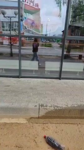 This video shows a person walking in front of a building with large glass windows. The scene appears to be an urban environment with a sidewalk and a street visible through the reflections in the glass.

**Subjects:**
*   **Person:** A single individual is walking from left to right across the frame, seen in reflection. They are wearing a dark-colored jacket and light-colored pants.
*   **Objects:** The building features large glass panels framed by dark metal. Inside the building, white benches are visible. The sidewalk is made of paving stones, and a dirt area with some sparse vegetation is in the foreground. Reflections show buildings, trees, and streetlights.
*   **Scene/Setting:** The scene is an outdoor urban setting. The glass panels suggest a modern building, possibly a public space, a bus stop shelter, or a commercial establishment. The reflections indicate a street with buildings and trees, hinting at a moderately developed area.

**Activity/Event:**
The primary activity is the person walking. The video captures their movement as they pass by the building.

**Notable Details:**
*   **Time of Day/Weather:** The lighting suggests it is daytime, possibly overcast or late afternoon, given the muted light and gray sky reflected in the glass.
*   **Location Cues:** The visible text "Пятница" (Friday) and "центр" (center) on a sign reflected in the window, combined with the general architecture and greenery, are consistent with a location in Russia. The overall environment appears somewhat modest rather than a bustling metropolis.
*   **Emotions:** No specific emotions are discernible from the footage. The scene is neutral and observational.

**Visible Text:**
The word "Пятница" (Friday) and "центр" (center) are visible, reversed, in the reflection on the glass.