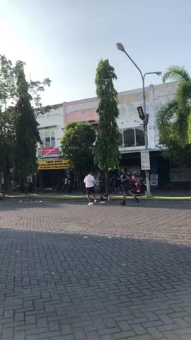 Two people are jogging on a paved area in front of a row of shops. The jogger in the foreground is wearing a white shirt and shorts, while the jogger behind them is wearing a dark shirt and shorts. Both are running away from the camera.

The shops appear to be on a street lined with trees. The buildings are modern with light-colored facades. A streetlamp is visible on the right side of the frame, and a palm tree is also present. The ground is paved with interlocking bricks. A sign with a red "30" is visible on the left side of the frame, indicating a speed limit. Another sign on the right has multiple icons, including one that appears to be a "no smoking" symbol.

The time of day appears to be morning or late afternoon, judging by the angle of the sunlight and the clear sky. The weather seems pleasant. The location is identified as Boro, East Java.

Visible text includes "TOKO KANA" on a yellow sign above one of the shops, and the "30" speed limit sign. Other text on signs is not clearly legible. The overall mood is one of an active, outdoor scene in a commercial area.