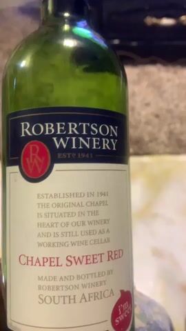 A person with dark skin and dark fingernails holds a green glass wine bottle. The bottle is labeled "ROBERTSON WINERY" at the top, with "EST? 1941" below it. A circular logo with "RW" is present. Further down, the label reads "CHAPEL SWEET RED," and "MADE AND BOTTLED BY ROBERTSON WINERY SOUTH AFRICA." A small, heart-shaped red sticker on the lower right of the label says "I'm sweet." The person's hand, wearing a ring on the middle finger, rotates the bottle slowly. The background is out of focus, showing a light-colored carpet or fabric and a dark, indistinct object.