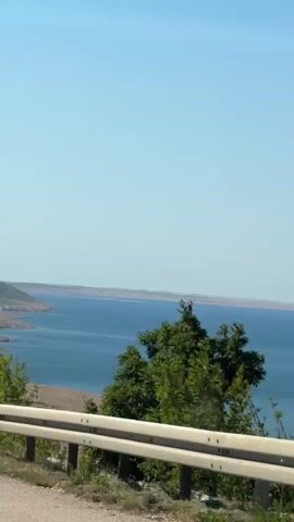 The video shows a view from a car, looking out towards a body of water and hills. The sky is clear and blue. There is a guardrail in the foreground, and trees on the left side of the frame. The car is moving, as indicated by the slight blur in the background.