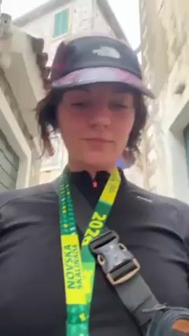 The video shows a woman walking down a narrow street. She is wearing a black long-sleeved shirt, a baseball cap with The North Face logo, and a medal with the words "NOVSKA SKALINADA 2023" on a green and yellow ribbon. She is facing the camera and smiling slightly. The street is paved with stones and lined with buildings. Other people are walking in the same direction. The lighting is overcast.