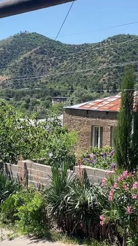 A video shows a hillside village with a stone house with a corrugated metal roof. Lush green trees and bushes are visible, along with some pink flowers. The sky is a clear blue. The video seems to be taken from a high vantage point, possibly a balcony or window, looking down onto the village. The sound consists of birds chirping.