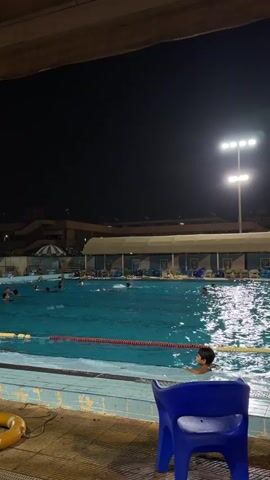 The video shows an outdoor swimming pool at night. Several people are swimming and playing in the water. There are lights illuminating the pool and surrounding area. In the foreground, a blue plastic chair and a yellow life preserver are visible. In the background, there are buildings and trees. The atmosphere appears to be recreational and lively.