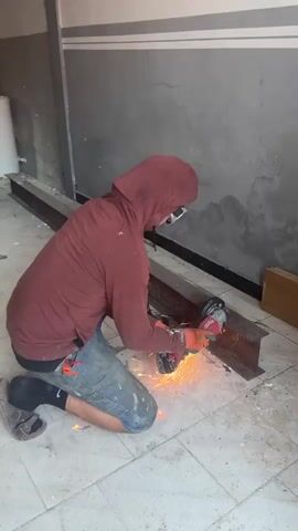 A person wearing a maroon hooded sweatshirt, denim shorts, black socks, safety glasses, and orange gloves is kneeling on a tiled floor. The person is using a red and silver angle grinder with a cutting disc to cut a piece of I-beam. Sparks are flying from the grinder as it cuts into the metal. The I-beam is resting on the floor. The background shows a grey wall with white horizontal stripes and some water stains.