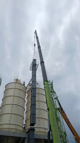 Two large industrial silos stand to the left of the frame. A tall, dark gray rectangular structure is being lifted by a crane. The crane boom is a dark gray color, and it extends from the right side of the frame. To the right of that crane, another crane boom, this one bright green, is visible. A person in a blue shirt and white hard hat is standing on a platform attached to the left silo, observing the lifting operation. The sky is overcast with gray clouds.