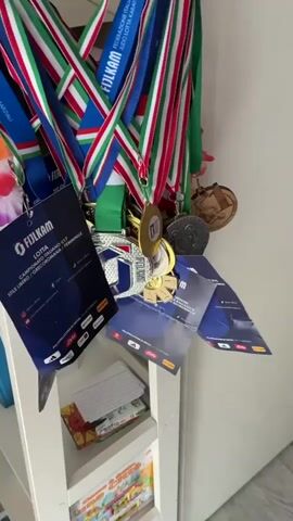 A hand reaches into the frame to adjust a collection of medals hanging on a white surface. The medals have colorful ribbons, predominantly in the Italian flag colors of green, white, and red, along with blue. Some of the medals are gold, silver, and bronze, and feature various insignia and text, including "FIJLKAM LOTTA CAMPIONATO ITALIANO U17". Several small paper cards are attached to the medals, displaying event details. In the background, on a white shelf, are some stacked papers and colorful children's books. The lighting suggests an indoor setting with natural light.