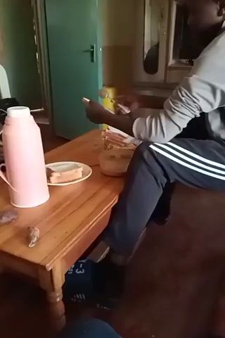 The video shows a person sitting at a wooden table, preparing food. The person is wearing a gray long-sleeved shirt and black pants with white stripes. On the table, there is a pink thermos, a plate with two slices of bread, a jar with a spread, and a knife. The person is using the knife to spread something from the jar onto a slice of bread. The room appears to be a living space, with a door and a cabinet visible in the background. The lighting is dim, suggesting it might be indoors or late in the day.
