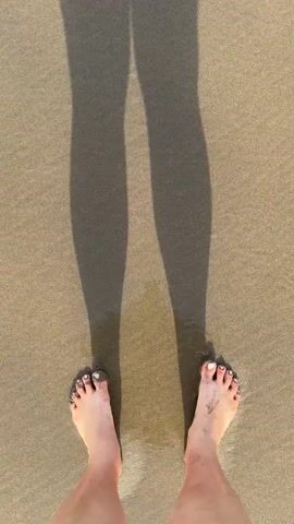 A top-down, close-up view of a person's bare feet on a sandy beach. The feet are dirty with sand. Long shadows of the person's legs are cast on the sand, indicating the sun is low in the sky. The person takes a step forward, and the edge of the water washes over their feet, creating ripples and small bubbles on the sand. The camera continues to track the feet as they move further into the shallow water.