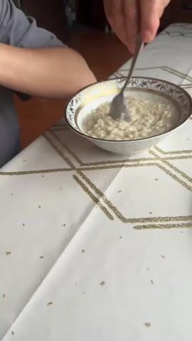 A person is sitting at a table with a bowl of oatmeal. They are stirring the oatmeal with a spoon. Another person is seen briefly reaching into the frame. The table is covered with a white tablecloth with a gold geometric pattern. The background is blurred but appears to be an indoor setting with wooden furniture.