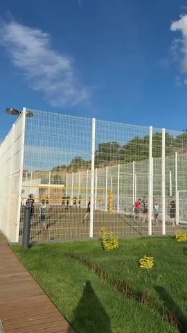 The video captures an outdoor volleyball court enclosed by a tall white mesh fence, illuminated by bright daylight under a clear blue sky with scattered clouds. A yellow volleyball net is stretched across the sand-colored court, where several individuals, dressed in athletic wear, are engaged in playing or preparing to play. In the foreground, a well-maintained grassy area with small green and yellow bushes separates the viewer from the court, alongside a wooden pathway on the left. A dark grey pole, possibly a light fixture, is also visible. Beyond the court, a natural hill covered with green trees forms the background. The scene depicts a lively recreational activity in a sunny, open-air environment.
