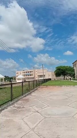 The video pans across a landscape featuring buildings, a fence, and a cloudy blue sky. In the foreground, a concrete walkway is visible, bordered by a black metal fence. Beyond the fence, a grassy area is apparent, interspersed with dirt patches. Some white structures with pyramidal roofs are present in the distance. Further back, larger buildings with multiple stories and windows are seen. A tall water tower with a tank on top stands to the right of the frame. Several individuals are walking along the walkway in the latter part of the video. The overall environment appears to be an outdoor campus or institutional setting.