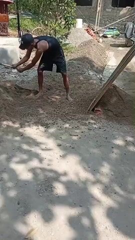 A man in a black tank top and black shorts, wearing a black baseball cap, is digging a pile of gravel with a shovel. He is barefoot. Behind him, another man in a pink shirt and blue shorts is using a shovel to move sand into a sieve. The sieve is propped up on a wooden frame, and sand is falling through it onto a pile below. In the background, there is a partially constructed building, a red cement mixer, and some trees. The scene appears to be an outdoor construction site.