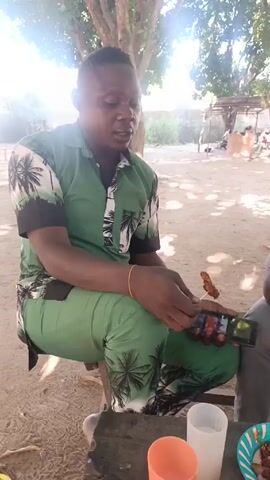 A man is sitting on a low bench outdoors, wearing a matching green patterned shirt and pants. He is holding a skewer with a piece of food in one hand and a mobile phone in the other. He appears to be looking at the phone while eating. In front of him on the bench are two plastic cups, one white and one orange, and a paper plate with food on it. Another person is sitting to his right, partially visible in the frame. The background includes trees providing shade, a sandy ground, and a distant building or structure.