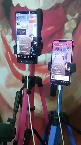 Two smartphones are mounted on separate tripods and are vertically oriented. Both phones are displaying what appears to be a live stream, possibly for social media or e-commerce. On the left, a larger, darker-colored phone shows a woman in the center of the screen, facing the camera and talking. The phone is charging, indicated by a white cable. The tripod holding this phone is pink and black. On the right, a lighter-colored smartphone, possibly an iPhone, also shows the same woman in a similar pose. This phone is also charging with a white cable, and its tripod is blue and black. The background is a patterned tapestry or fabric with red, pink, and cream colors, featuring what looks like a stylized face or figure. The lighting suggests an indoor setting. The overall setup indicates someone is likely engaged in live streaming, possibly demonstrating a product or interacting with an audience.