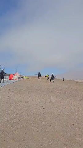 The video shows a group of people preparing for paragliding on a sandy hill under a sky with a mix of clouds and blue patches. Five individuals are visible. One person is standing on the left side, wearing a dark jacket, dark pants, and a hat. They have a backpack on. Three individuals are closer to the center of the frame. One wears a yellow jacket and a helmet. Another wears a gray jacket with a large backpack and a helmet. They appear to be untangling lines in preparation for a flight. The fifth person, possibly a child, is further down the hill. Several colorful paragliding wings are laid out on the ground. The terrain appears to be sandy or gravelly, with a mountainous landscape visible in the distance. The overall setting suggests an outdoor recreational activity in a dry, elevated environment.