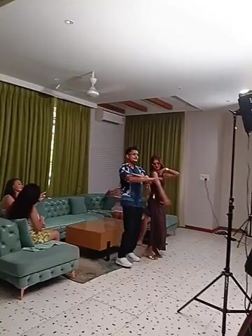 A medium shot shows a living room setting with a man and a woman practicing dance moves while others watch. The room is well-lit with natural light from large windows covered by green drapes. The floor is decorated with speckled tiles. A light turquoise sectional couch sits on the left with three people lounging on it. A wooden coffee table sits in front of the couch on top of a rug. The man, wearing a blue and white patterned shirt, black pants, and white shoes, and the woman, wearing a brown dress and gold sandals, are in the center of the room. A light stand with a large light fixture is to the right, close to a white door. A few air conditioning units can be seen mounted on the wall. The general atmosphere appears relaxed.