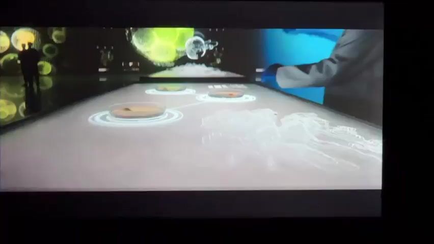 The video opens with a wide shot of a futuristic-looking exhibition space. A long, illuminated table dominates the foreground, with various circular displays on its surface. In the background, a large screen displays vibrant, animated graphics, including what appears to be cellular structures and a DNA helix.

As the video progresses, a person in a lab coat is seen interacting with the display on the large screen, touching it with their gloved hands. Another person, silhouetted against the bright graphics, stands to the left of the table, appearing to observe the display.

The illuminated table in the foreground projects holographic-like images, including what looks like a wireframe of an anatomical structure. The animated graphics on the background screen shift and change, at one point showing a swirling mass of red particles that coalesce into the DNA helix.

Towards the end of the clip, the animated graphics on the table become more prominent, with a large, white, three-dimensional object, resembling a flower or a cloud-like formation, emerging from the surface. The lab-coated individual continues to interact with the display, their hands positioned over the table. The overall environment suggests a high-tech science or research exhibition, with a focus on biotechnology or genetics.