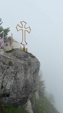 A vertical video shows a golden cross on top of a rocky cliff. The cross has a distinctive angular design. In the background, fog obscures the view of the surrounding landscape, but hints of trees and greenery can be seen on the slope below. A railing and a small structure are partially visible on the left side of the cliff, along with two figures wearing pink tops, suggesting a viewing platform or tourist area. The overall atmosphere is serene and slightly mysterious due to the fog.