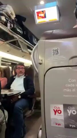 The camera moves slowly through the inside of a passenger train cabin. Seated passengers are visible in the background, and there is luggage overhead. The back of a seat is prominent in the foreground. It has an advertisement for Club Yo iryo written in Spanish along with a QR code. A small monitor is mounted overhead.