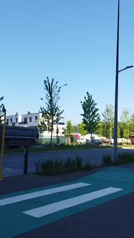 The video captures a street scene with a clear blue sky. In the foreground, a pedestrian crossing is marked with green and white stripes on the asphalt. To the left, a black pole stands vertically. Beyond the street, there are trees and buildings. A red truck with a yellow trailer is visible on the street, driving away from the camera. A white car is also seen on the street, driving in the same direction. In the background, there are modern-looking buildings with white facades and dark-framed windows, surrounded by greenery and trees. A tall lamppost with a light fixture stands on the right side of the frame. The overall impression is of a suburban or newly developed area.