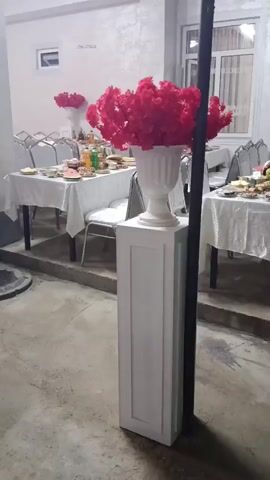The video shows a dimly lit outdoor area, possibly a patio or courtyard, set up for a gathering. Two long tables are covered with white tablecloths and laden with food and drinks, suggesting a meal or a party. Several chairs are arranged around the tables. In the foreground, a white pedestal stands with a large white urn overflowing with bright red artificial flowers. To the right of the pedestal, a black pole is visible. A man wearing a black shirt, dark pants, and a light-colored cap walks into the frame from the right, pauses briefly, and then walks out of frame to the right. He appears to be wiping his face or adjusting something on his face. The background features windows with sheer curtains and what appears to be a wall with textured wallpaper. The overall impression is a pre-event setup or a lull during a gathering.